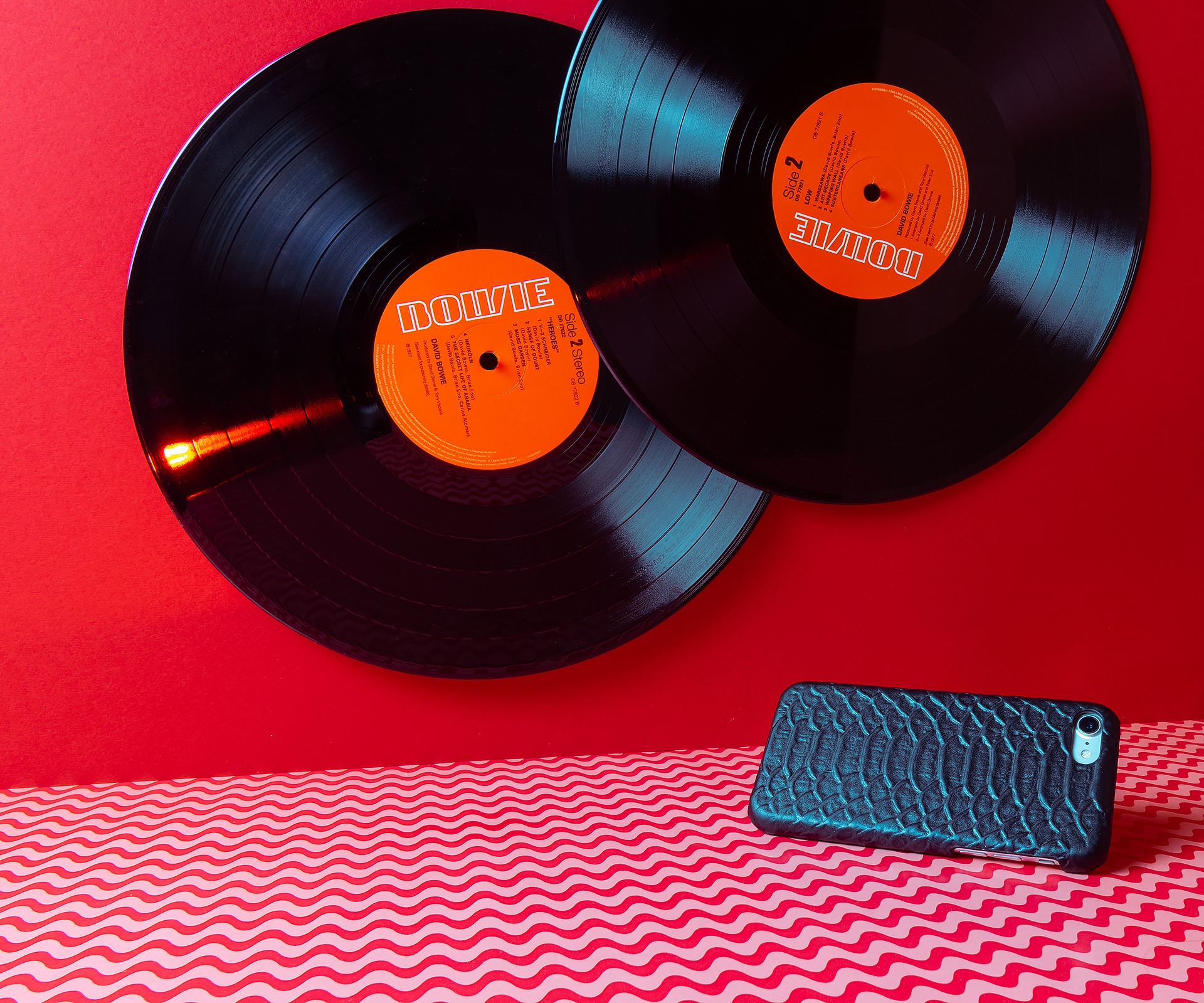 Dwie płyty winylowe Davida Bowie z okładkami albumu 'Heroes' na czerwonym tle i pokrowiec na telefon z wzorem skóry węża na czerwono-białym falistym tle.