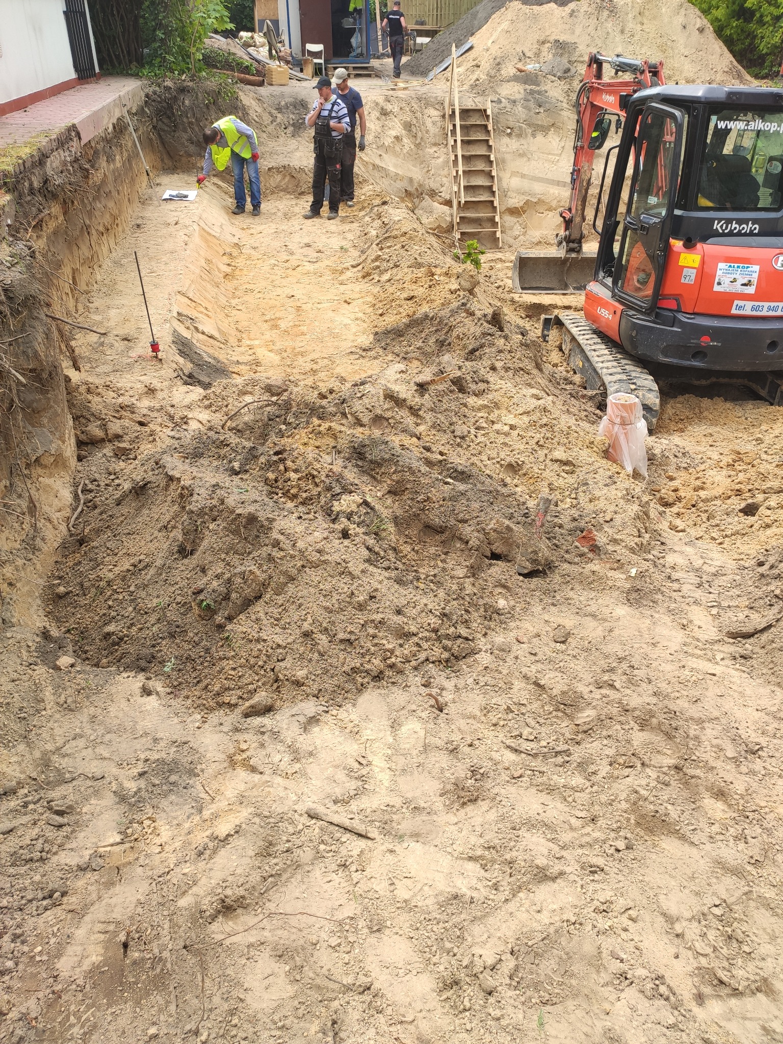 Wykop pod fundamenty z widoczną mini koparką Kubota U55-4, pracownikami mierzącymi głębokość wykopu oraz schodami ziemnymi prowadzącymi na górę.