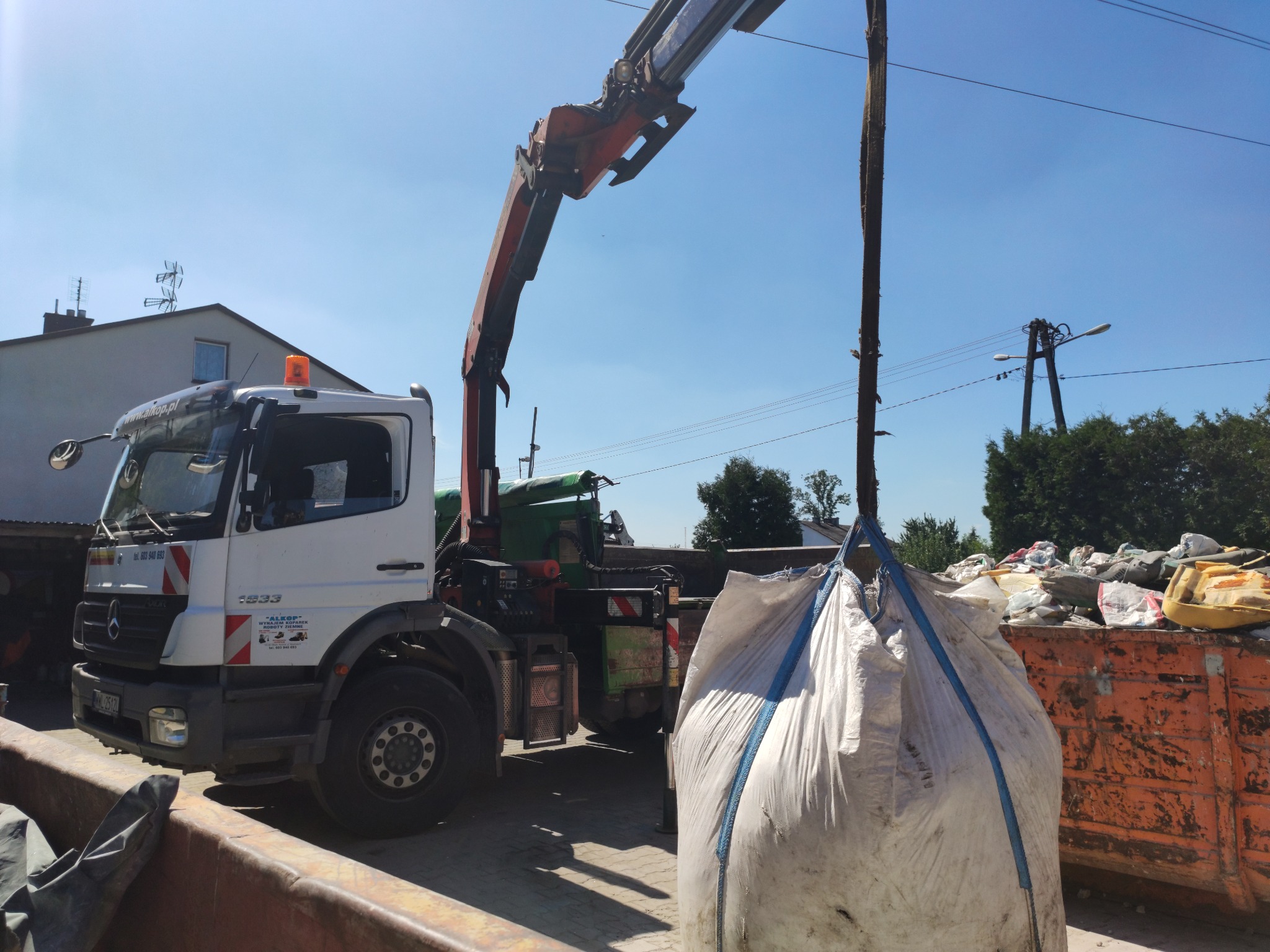 Ciężarówka z dźwigiem podnosi duży, biały worek z zawartością nad pomarańczowym kontenerem wypełnionym odpadami budowlanymi na tle domów i drzew.