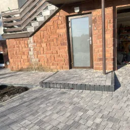 Ułożona szara kostka brukowa prowadząca do wejścia domu z ceglaną elewacją i schodami. Widoczny fragment rabaty z ziemią.