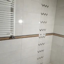 Wykończona łazienka z białymi płytkami, dekoracyjnym pasem z motywem jodełki i grzejnikiem drabinkowym z chromowanym zaworem.