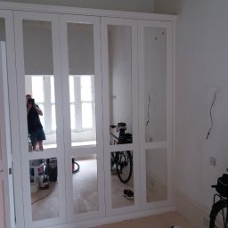 Perfect P - Biała szafa wnękowa z lustrami w trakcie montażu, widoczne niedociągnięcia wykończeniowe na ścianie i podłodze, odbicie osoby robiącej zdjęcie w lustrze, oraz rowery i buty w pomieszczeniu.