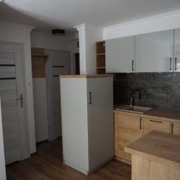 Perfect P - Nowoczesna kuchnia z białymi szafkami górnymi, drewnianymi szafkami dolnymi, blatem imitującym drewno i dekoracyjnymi płytkami na ścianie nad blatem. Widoczne drzwi w tle.