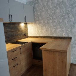 Perfect P - Fragment nowej kuchni z drewnianymi szafkami, czarnymi uchwytami i indukcyjną płytą grzewczą, na ścianie tapeta w liście, widoczny piekarnik z wyświetlaczem.