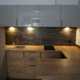 Nowoczesna kuchnia z szarymi szafkami lakierowanymi, drewnianym blatem i płytkami z motywem roślinnym, czarny zlew i płyta indukcyjna.