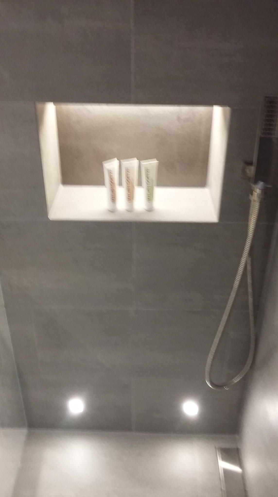 Wnęka w szarej, wykafelkowanej ścianie kabiny prysznicowej w Pruszkowie, z trzema tubkami kosmetyków i słuchawką prysznicową po prawej stronie.
