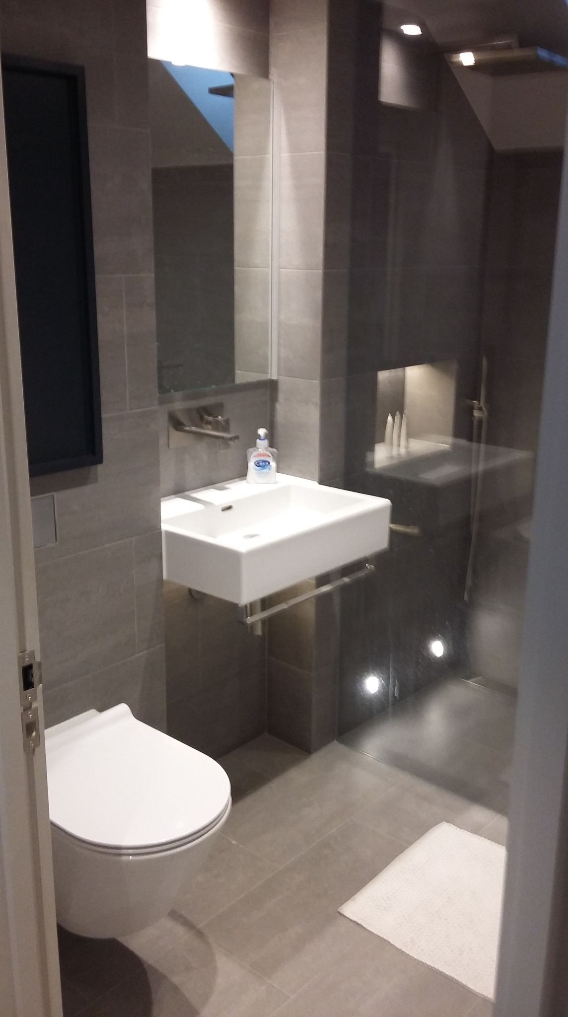 Nowoczesna łazienka w odcieniach szarości z podwieszaną toaletą, umywalką z baterią ścienną i kabiną prysznicową typu walk-in z oświetleniem punktowym.