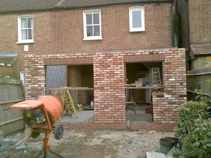 Budowa nowego, ceglanego rozszerzenia domu z widocznym mieszalnikiem do betonu na pierwszym planie i oknami na górze