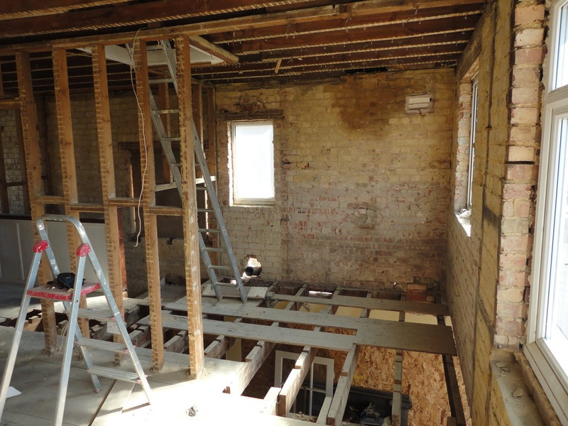 Wnętrze w trakcie demontażu: odsłonięta cegła, drewniane legary podłogowe z widoczną przestrzenią poniżej, dwie aluminiowe drabiny, małe okno w ścianie, surowe drewno stropu.