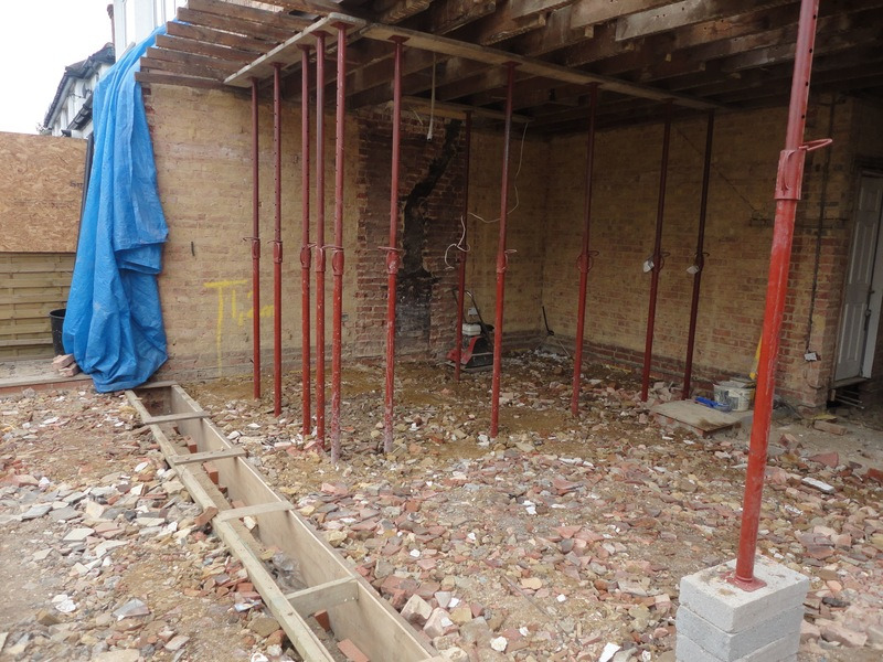 Wnętrze w trakcie przebudowy: podparte belki stropowe, odsłonięte cegły, gruz i drewniane szalunki pod fundamenty.