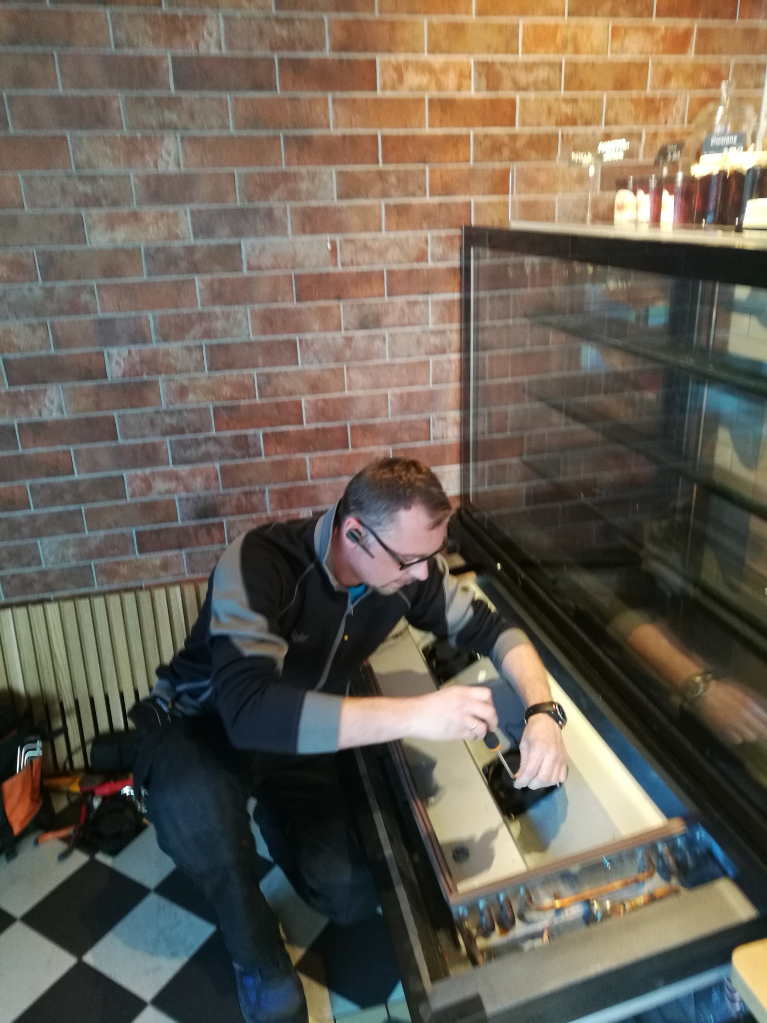 Mężczyzna w okularach i słuchawce Bluetooth naprawia chłodniczą witrynę sklepową za pomocą śrubokręta; widoczne miedziane rurki i wentylator; w tle ściana z cegły i półka z butelkami.