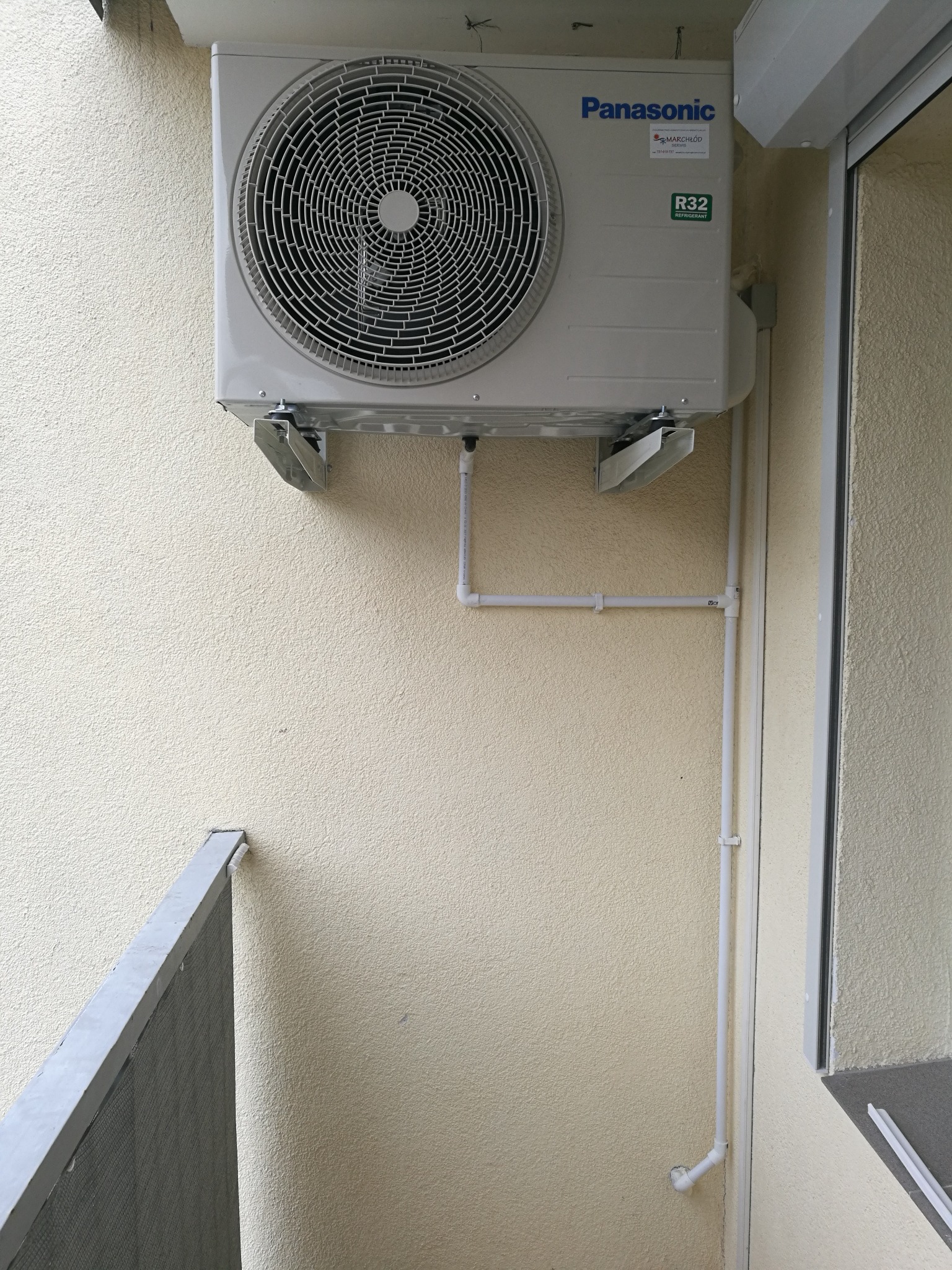 Zewnętrzna jednostka klimatyzacji Panasonic zamontowana na ścianie budynku z widocznymi rurami odprowadzającymi skropliny.