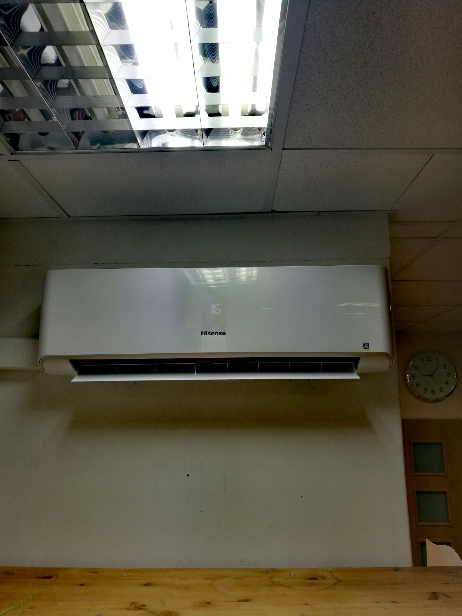 Biały klimatyzator Hisense zamontowany na ścianie pod sufitem z widoczną kratką oświetleniową i zegarem analogowym.