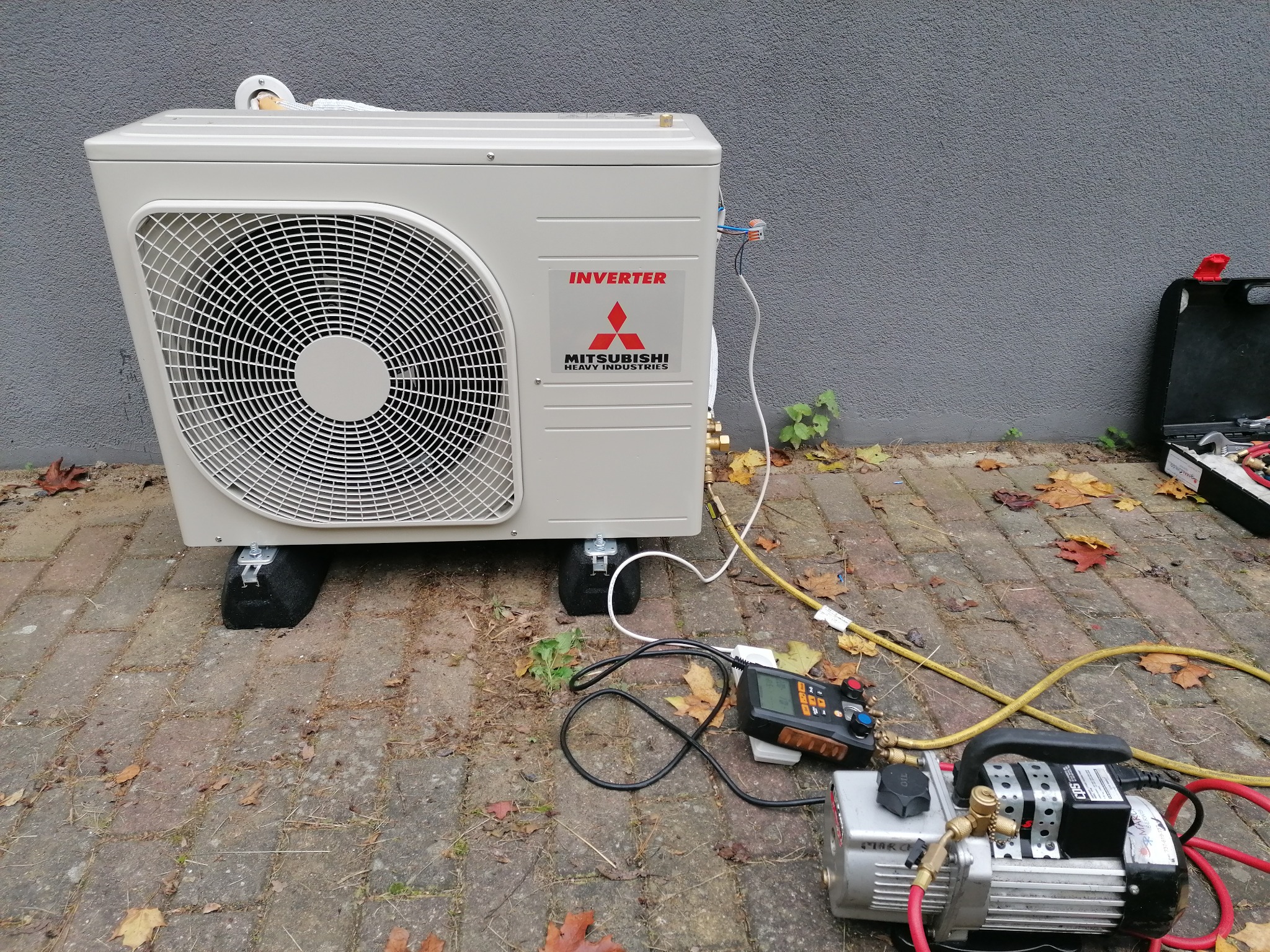 Zewnętrzna jednostka klimatyzatora Mitsubishi Heavy Industries, podłączona do pompy próżniowej i manometru cyfrowego, ustawiona na czarnych podkładkach antywibracyjnych, na tle szarej ściany...