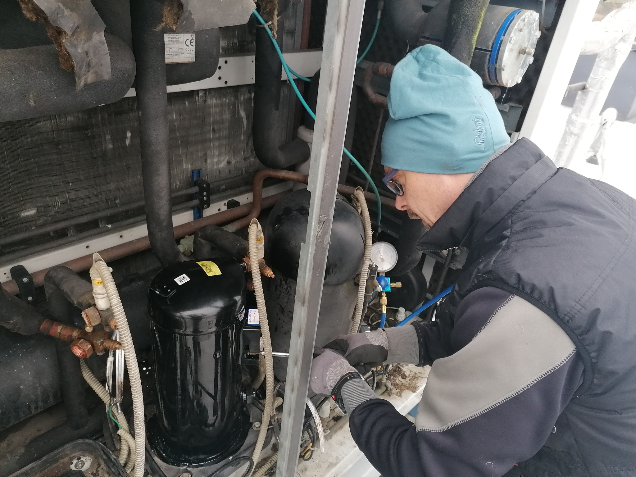 Mężczyzna w czapce i okularach serwisuje sprężarkę chłodniczą, widoczne rury, manometry i przewody.