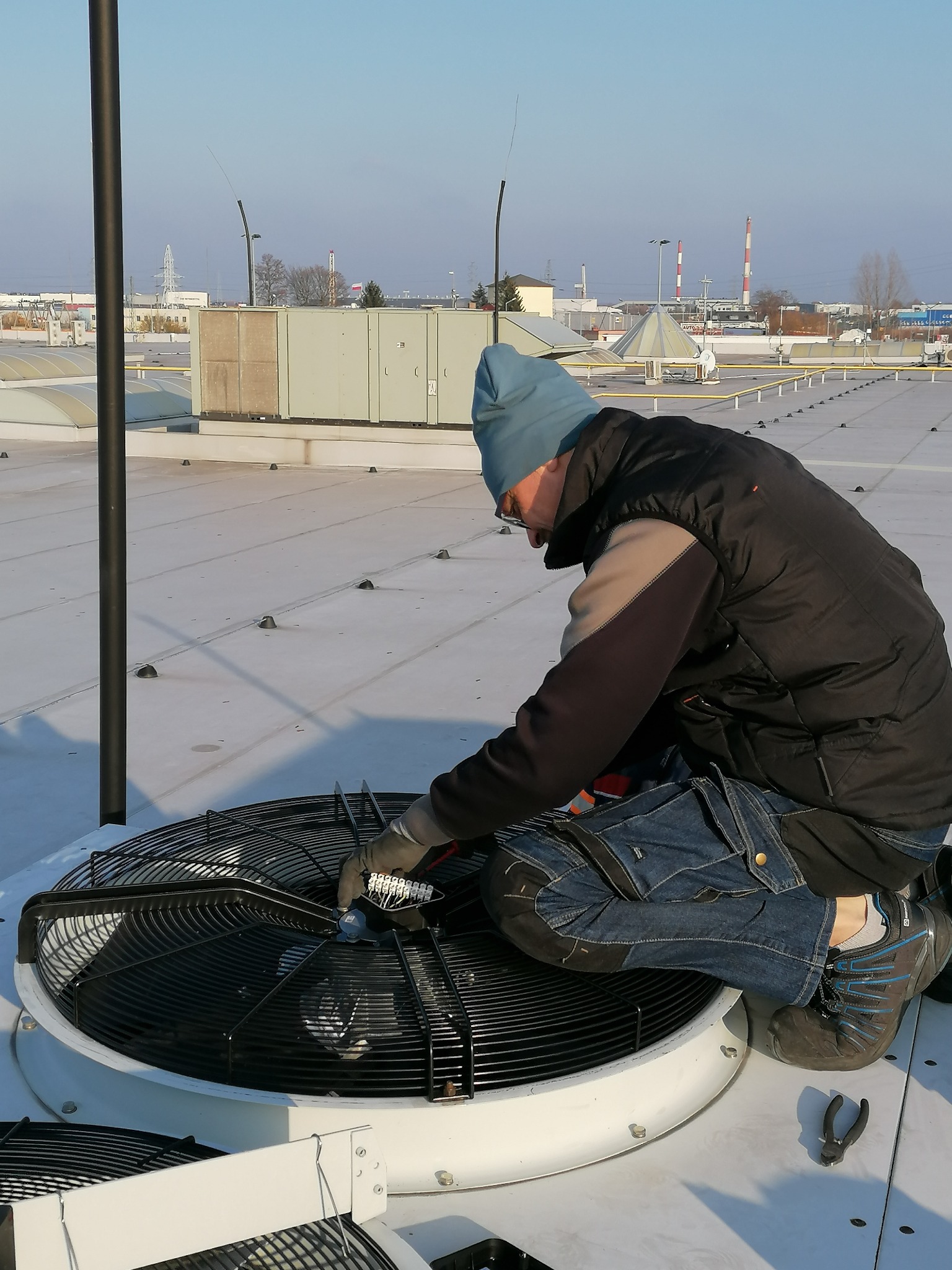 Pracownik w czapce i kamizelce, klęczy na dachu budynku, dokonując regulacji wentylatora w jednostce zewnętrznej klimatyzacji, widoczne narzędzia, w tle zabudowania przemysłowe