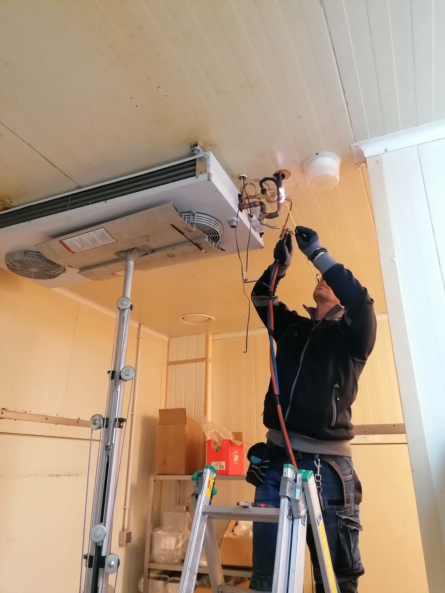 Instalator na drabinie lutuje rury do chłodnicy sufitowej w pomieszczeniu chłodniczym, widoczne narzędzia i przewody.