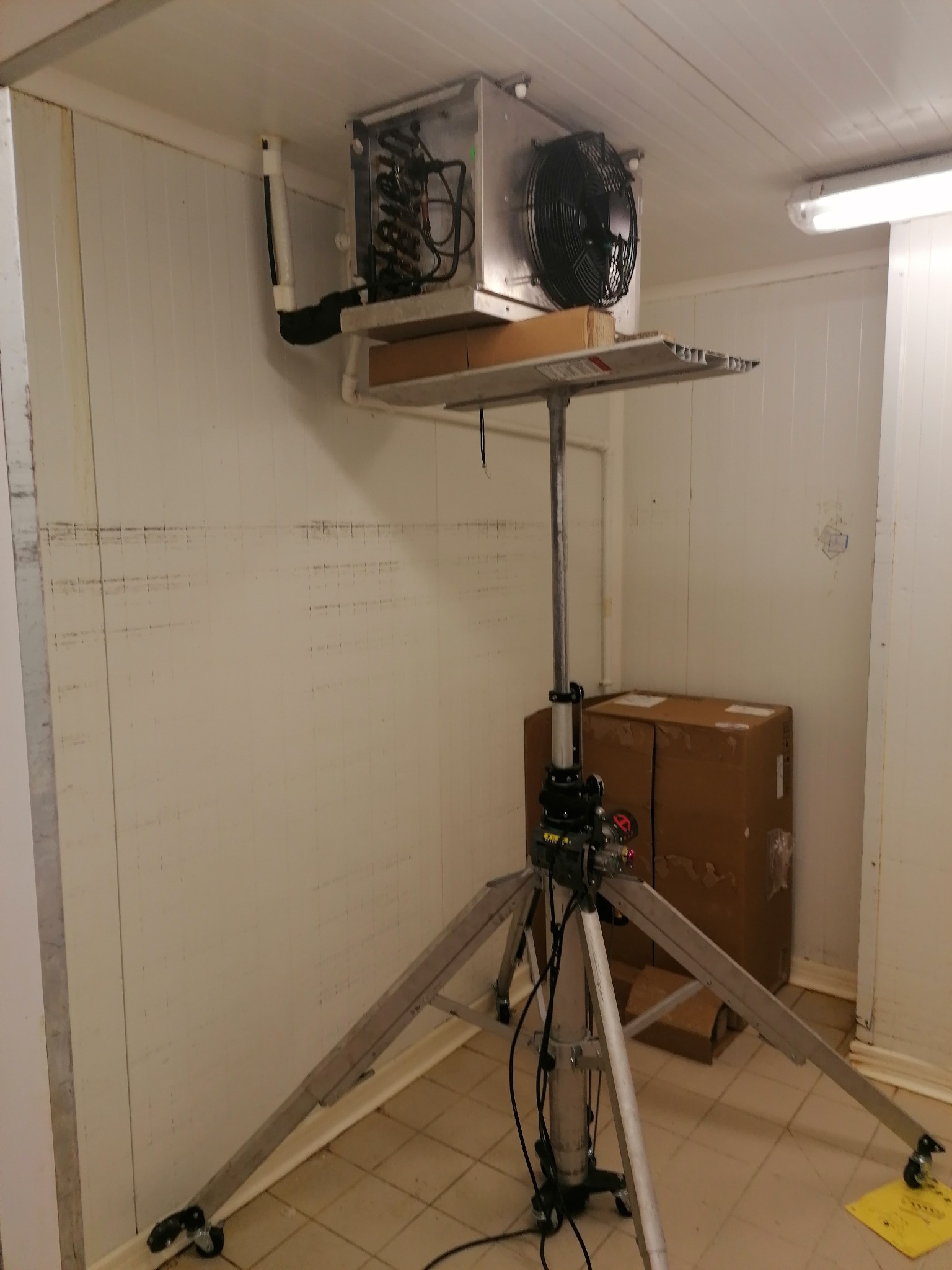 Wnętrze chłodni z zamontowanym na metalowym statywie agregatem chłodniczym, widoczne przewody i wentylator