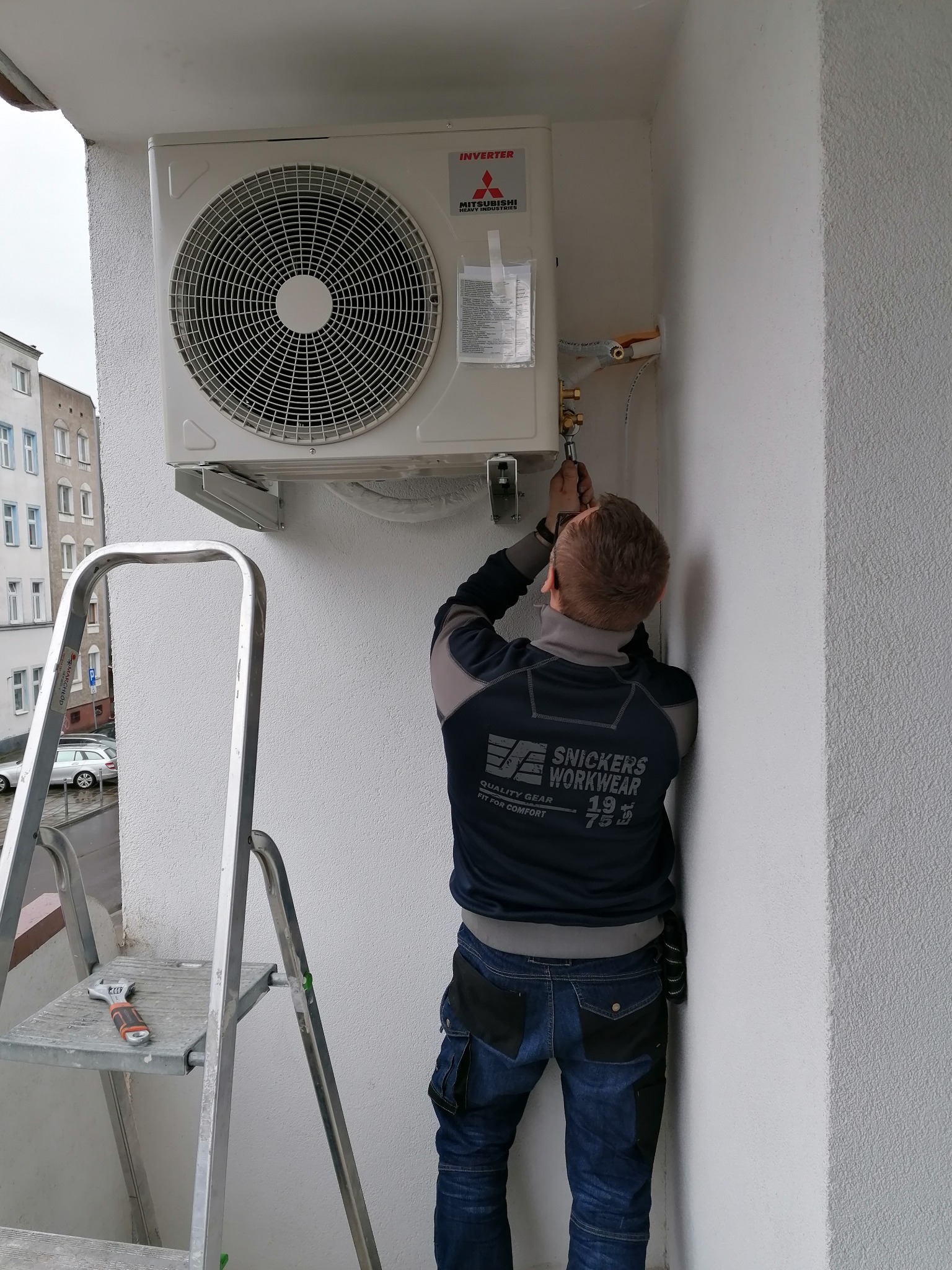 Instalator podłącza zewnętrzną jednostkę klimatyzacyjną Mitsubishi na balkonie, używając klucza nastawnego; drabina i narzędzia obok.