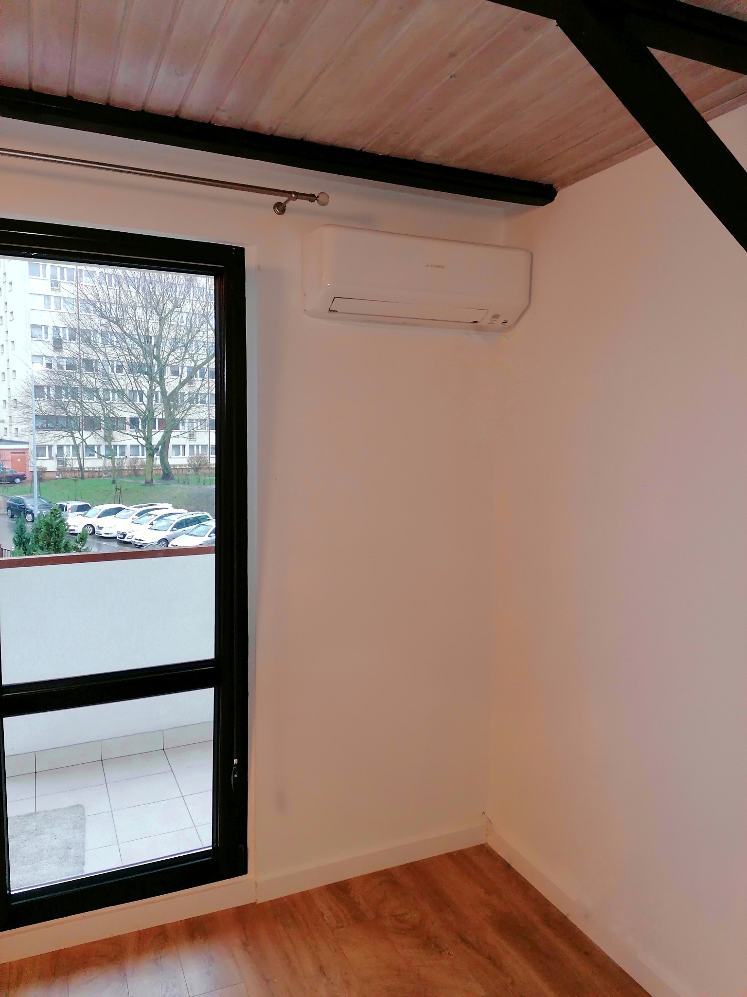 Biały klimatyzator zamontowany na ścianie w pomieszczeniu w Szczecinie, widok z okna na parking i blok mieszkalny, drewniana podłoga i sufit.