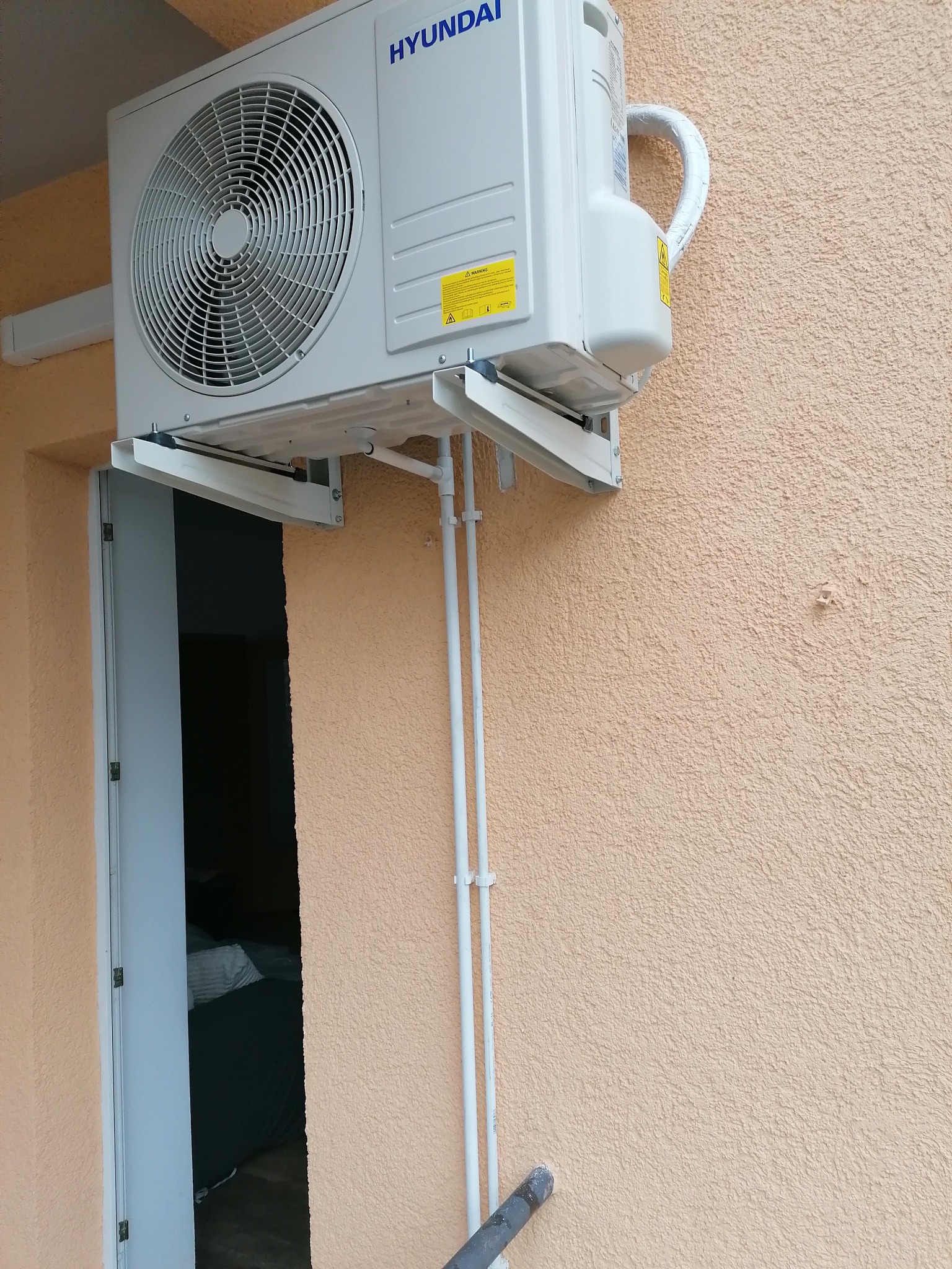 Zewnętrzna jednostka klimatyzacyjna marki Hyundai zamontowana na elewacji budynku obok otwartych drzwi.