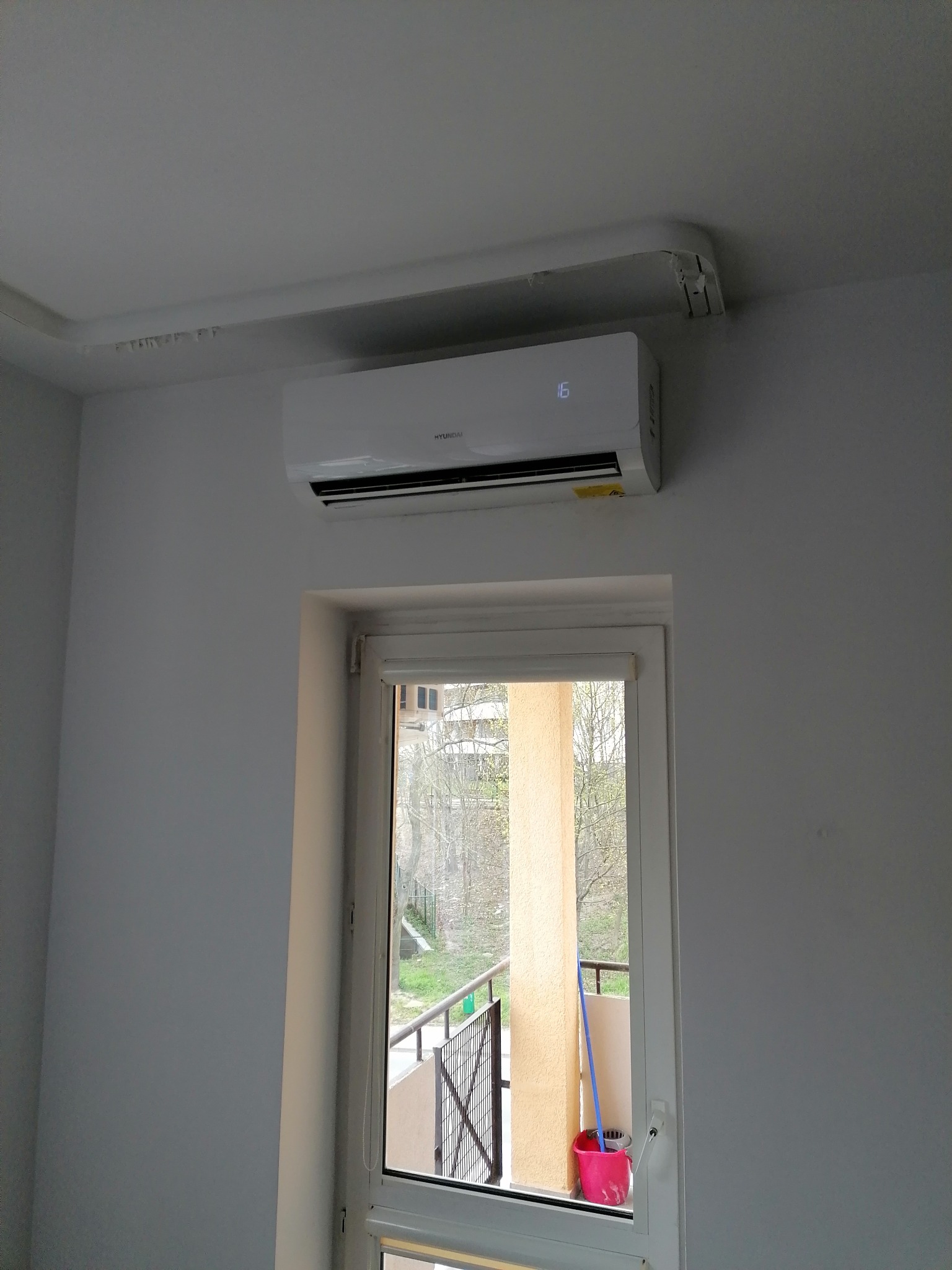 Biały klimatyzator Hyundai zamontowany na ścianie nad oknem, widoczna instalacja z rurami na suficie, balkon za oknem z widocznym wiadrem i mopem.