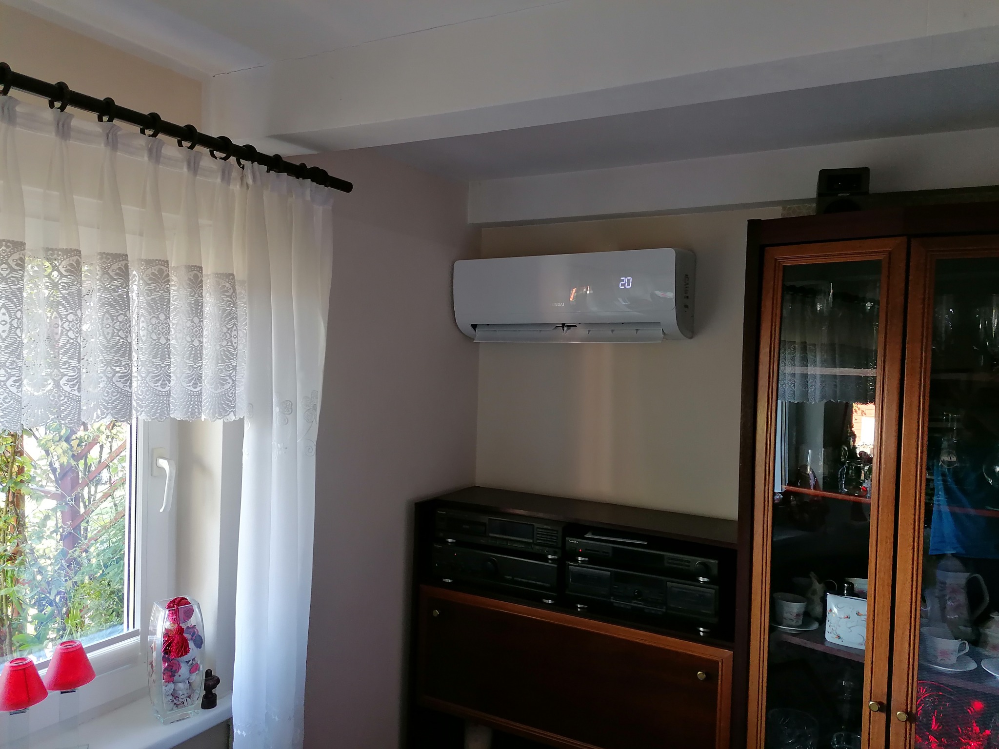 Biały klimatyzator ścienny z wyświetlaczem temperatury zamontowany w pokoju z oknem, zasłonami, szafką i sprzętem audio.