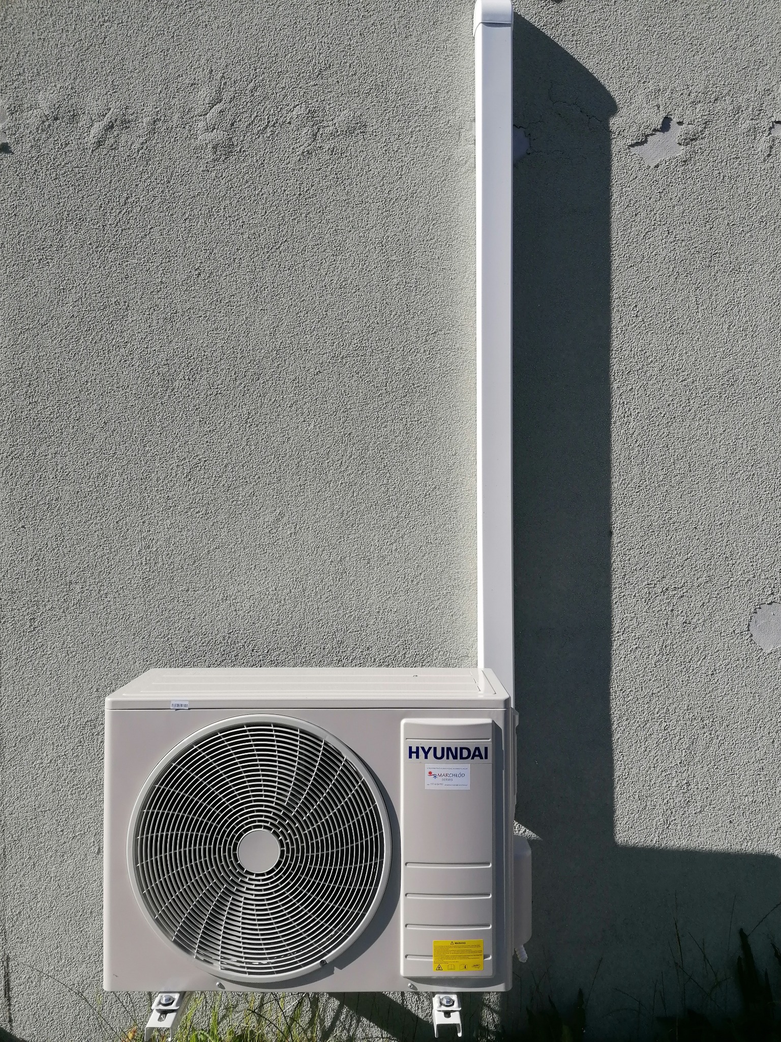 Zewnętrzna jednostka klimatyzatora marki Hyundai zamontowana na szarej, teksturowanej ścianie budynku z widoczną białą rurą odprowadzającą skropliny.