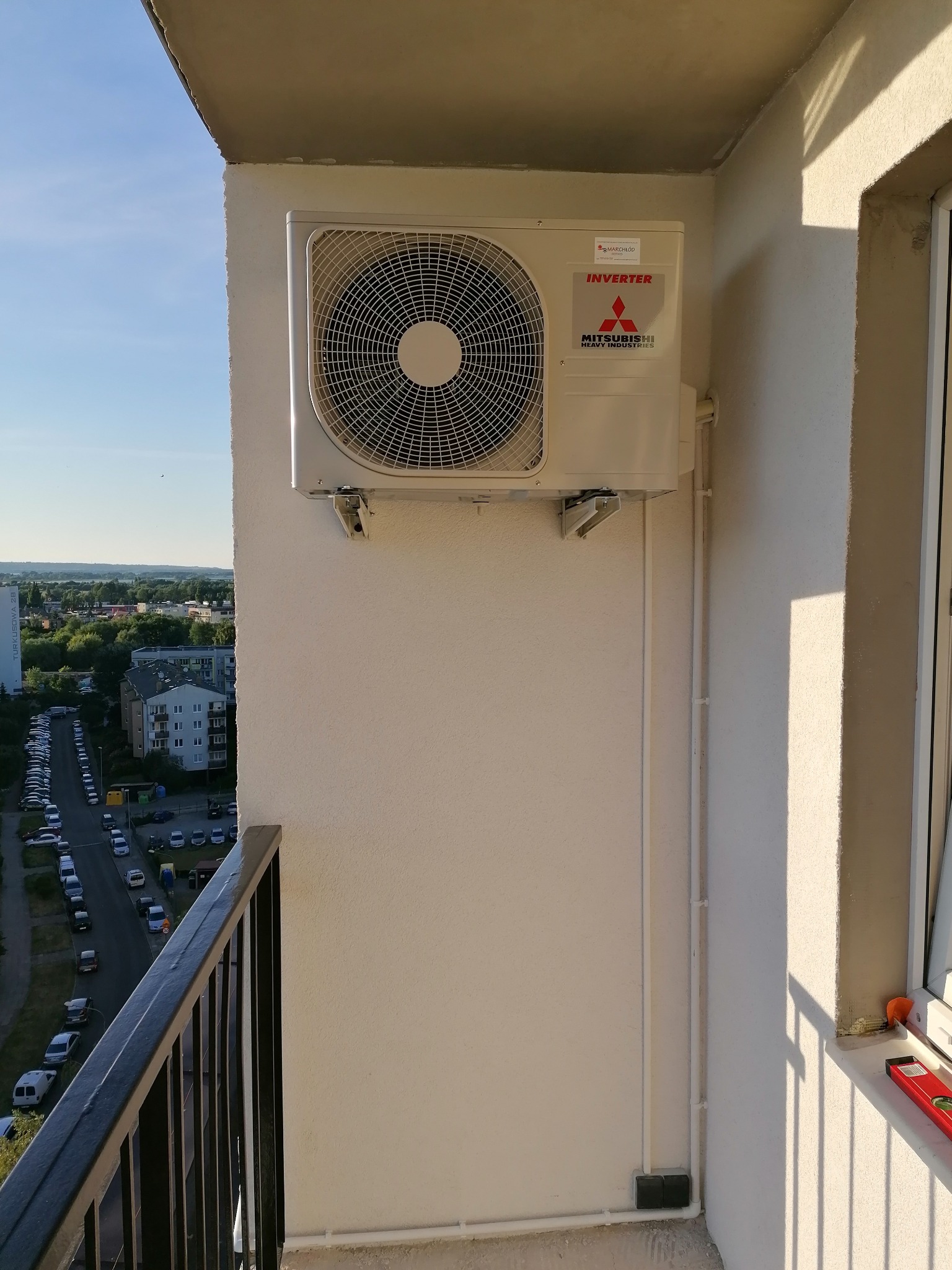 Klimatyzator Mitsubishi Inverter zamontowany na balkonie z widokiem na miasto, widoczne rury instalacyjne i poziomica na parapecie.