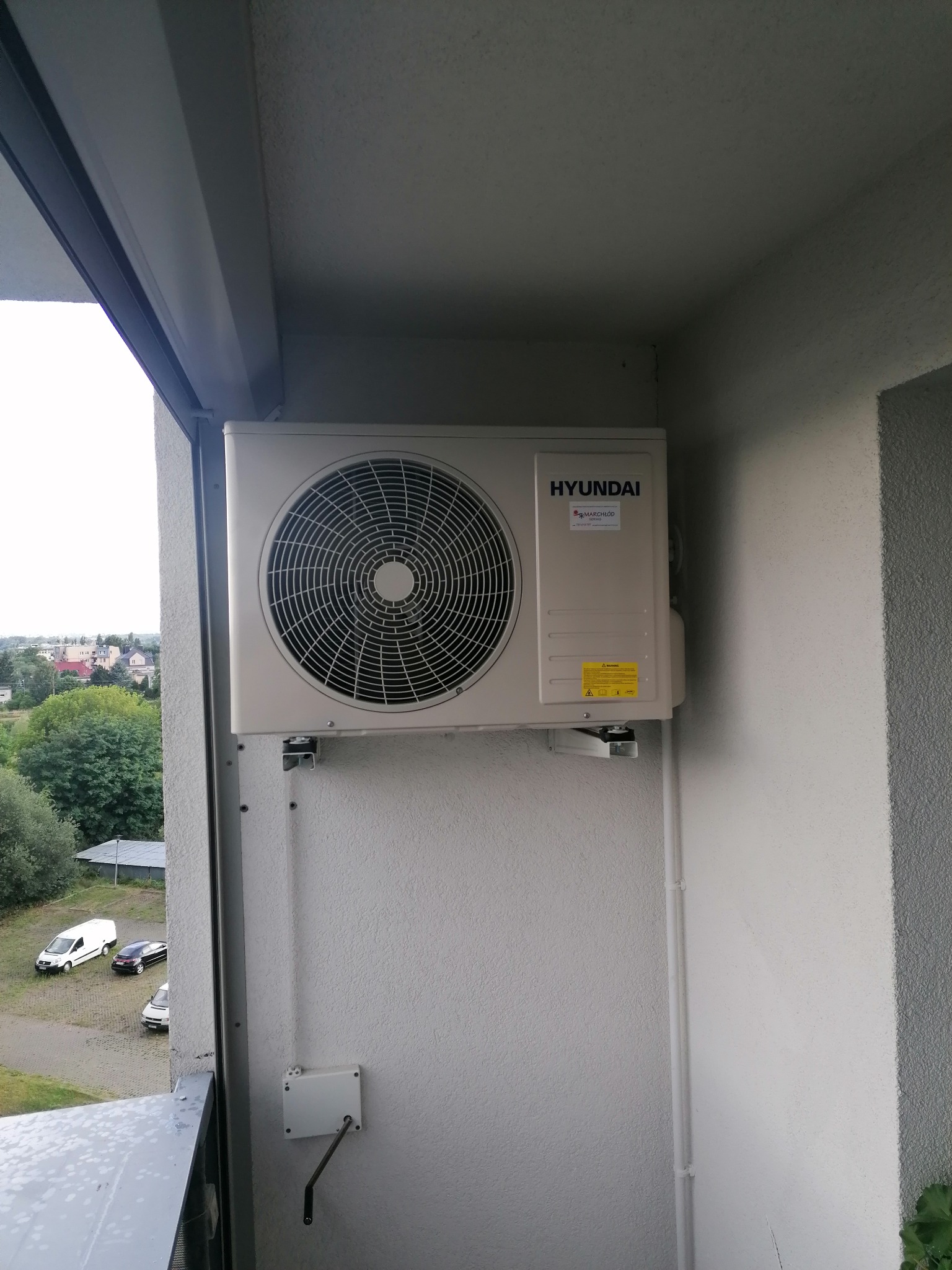 Zewnętrzna jednostka klimatyzatora Hyundai zamontowana na balkonie z widokiem na drzewa i budynki w oddali.