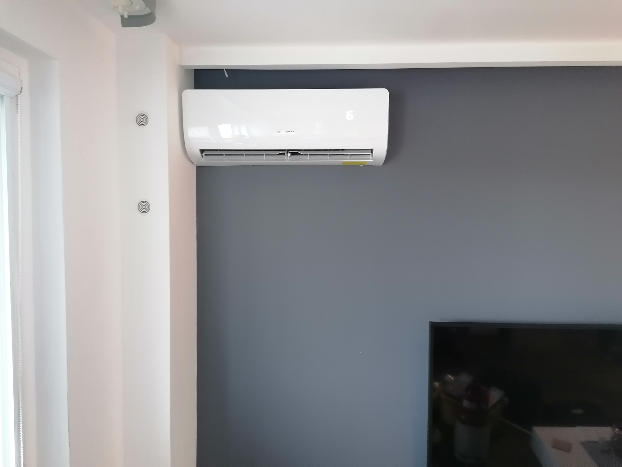 Biały klimatyzator ścienny na szarej ścianie w nowoczesnym wnętrzu, widoczny fragment telewizora w rogu.