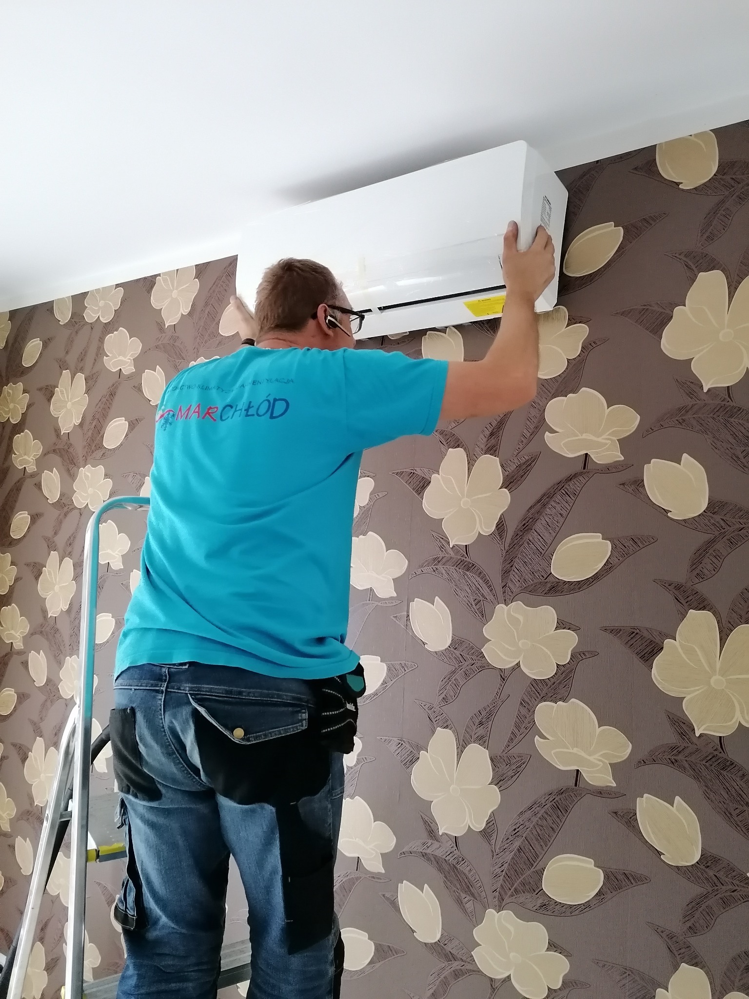 Mężczyzna na drabinie montuje białą jednostkę wewnętrzną na ścianie z tapetą w kwiaty, widoczny tył niebieskiej koszulki z logo firmy.