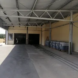 Wnętrze hali produkcyjnej z metalową konstrukcją dachu, betonową posadzką, bramami wjazdowymi i rzędem zbiorników IBC na paletach wzdłuż ściany.