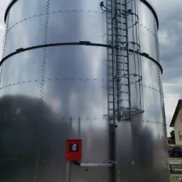 Srebrzysty silos zbożowy z zewnętrzną klatką schodową, zamontowaną czerwoną skrzynką kontrolną i fragmentem budynku w tle.