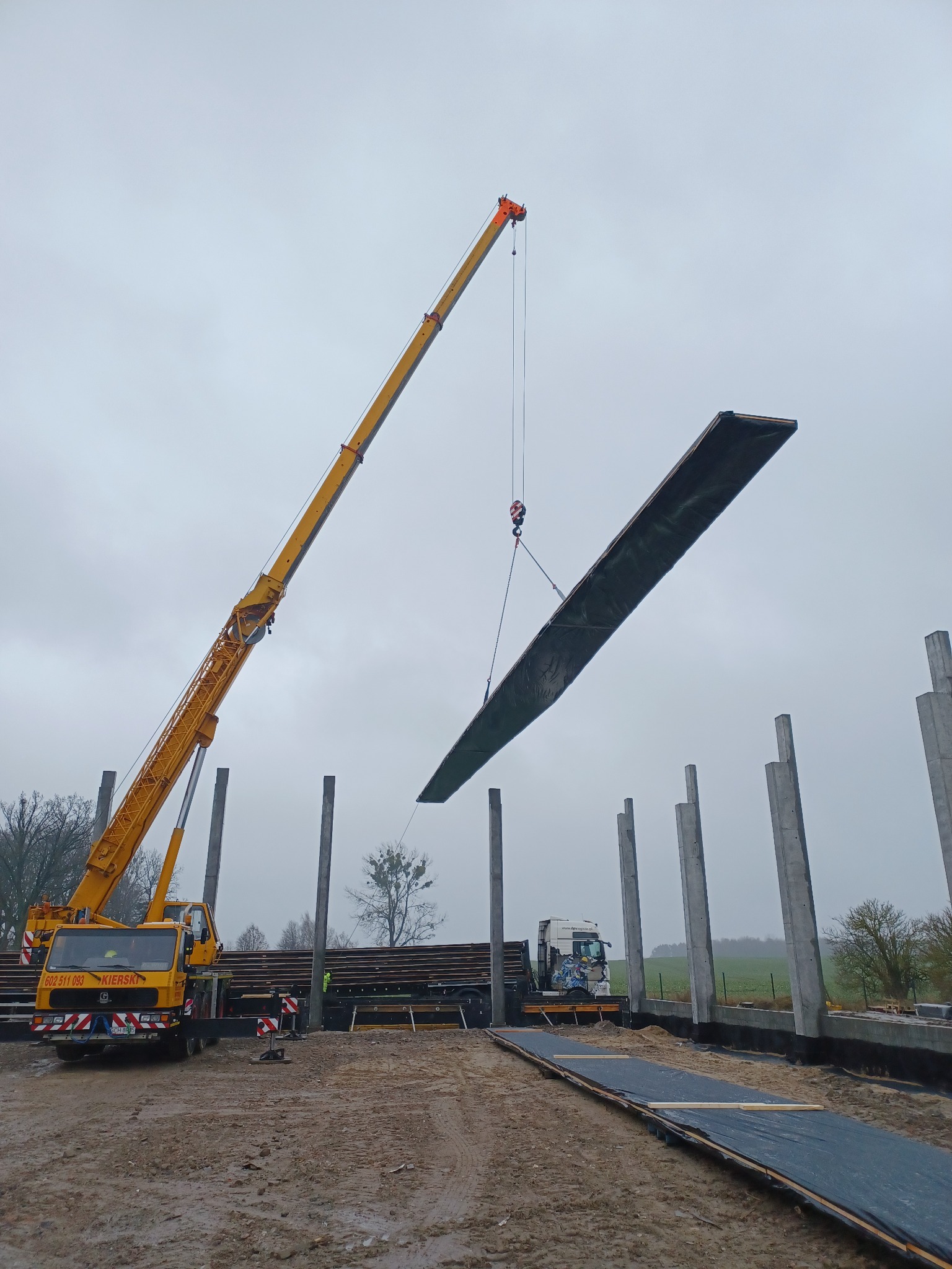 Żółty dźwig podnosi betonowy element konstrukcyjny na budowie, widoczne betonowe słupy i teren budowy w pochmurny dzień.