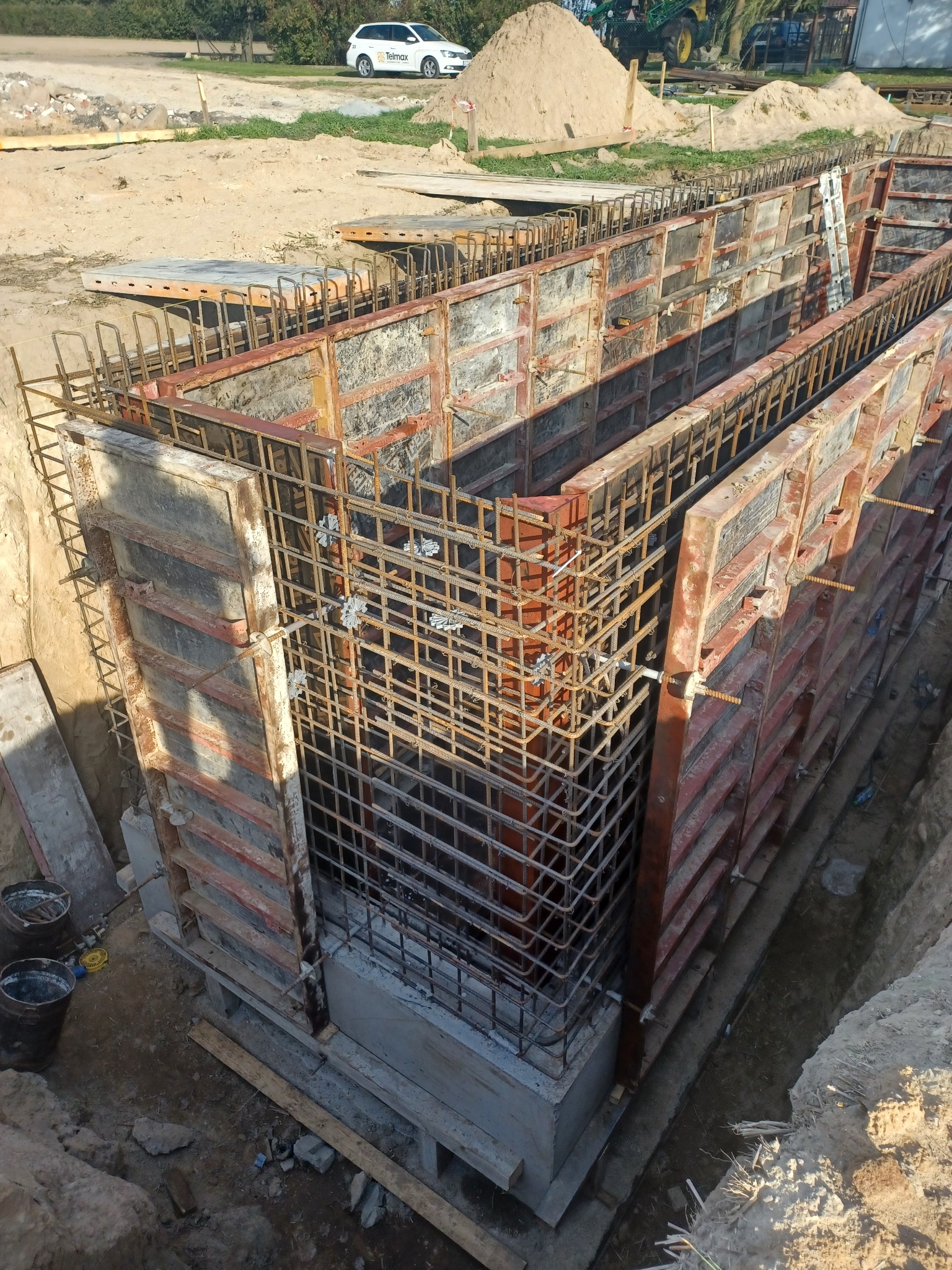 Szalunek zbrojeniowy przygotowany pod wylanie fundamentów, widoczne stalowe pręty i deski szalunkowe, na drugim planie hałda piasku i sprzęt budowlany.
