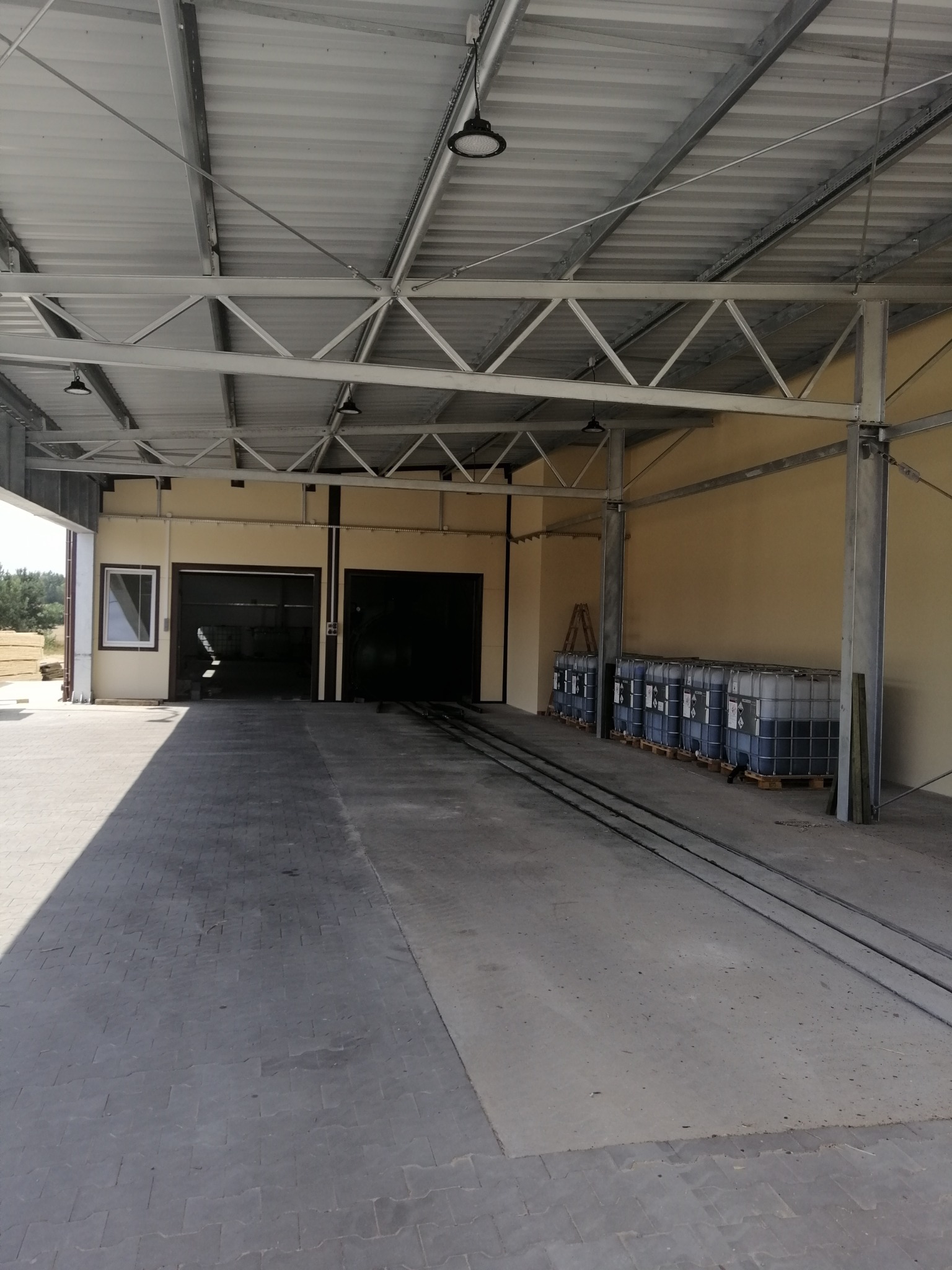 Wnętrze hali produkcyjnej z metalową konstrukcją dachu, betonową posadzką, bramami wjazdowymi i rzędem zbiorników IBC na paletach wzdłuż ściany.