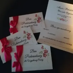 Karta podarunkowa na masaż i rehabilitację z różową wstążką i kwiatowym motywem, obok bon upominkowy z adresem gabinetu.