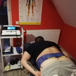 Pacjent leży na brzuchu na łóżku zabiegowym z elektrodami na plecach, podłączonymi do urządzenia do elektrostymulacji; na ścianie plakat z podstawowymi badaniami neurologicznymi.