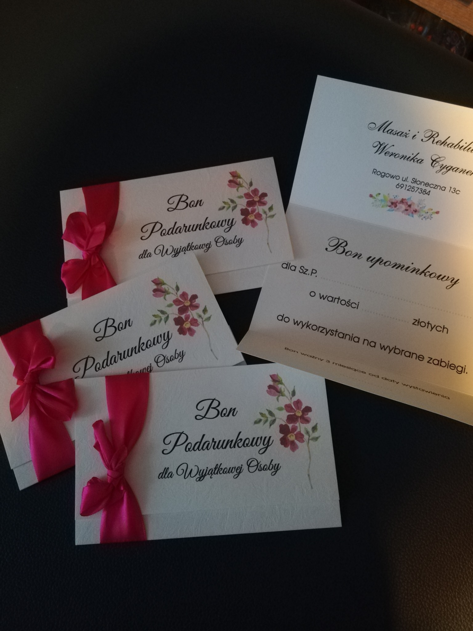 Karta podarunkowa na masaż i rehabilitację z różową wstążką i kwiatowym motywem, obok bon upominkowy z adresem gabinetu.