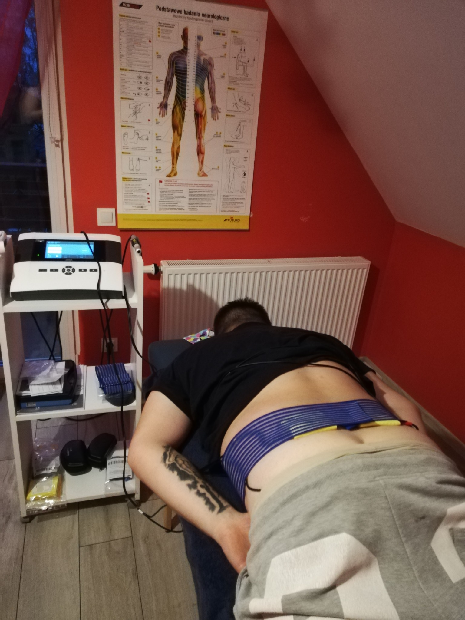 Pacjent leży na brzuchu na łóżku zabiegowym z elektrodami na plecach, podłączonymi do urządzenia do elektrostymulacji; na ścianie plakat z podstawowymi badaniami neurologicznymi.