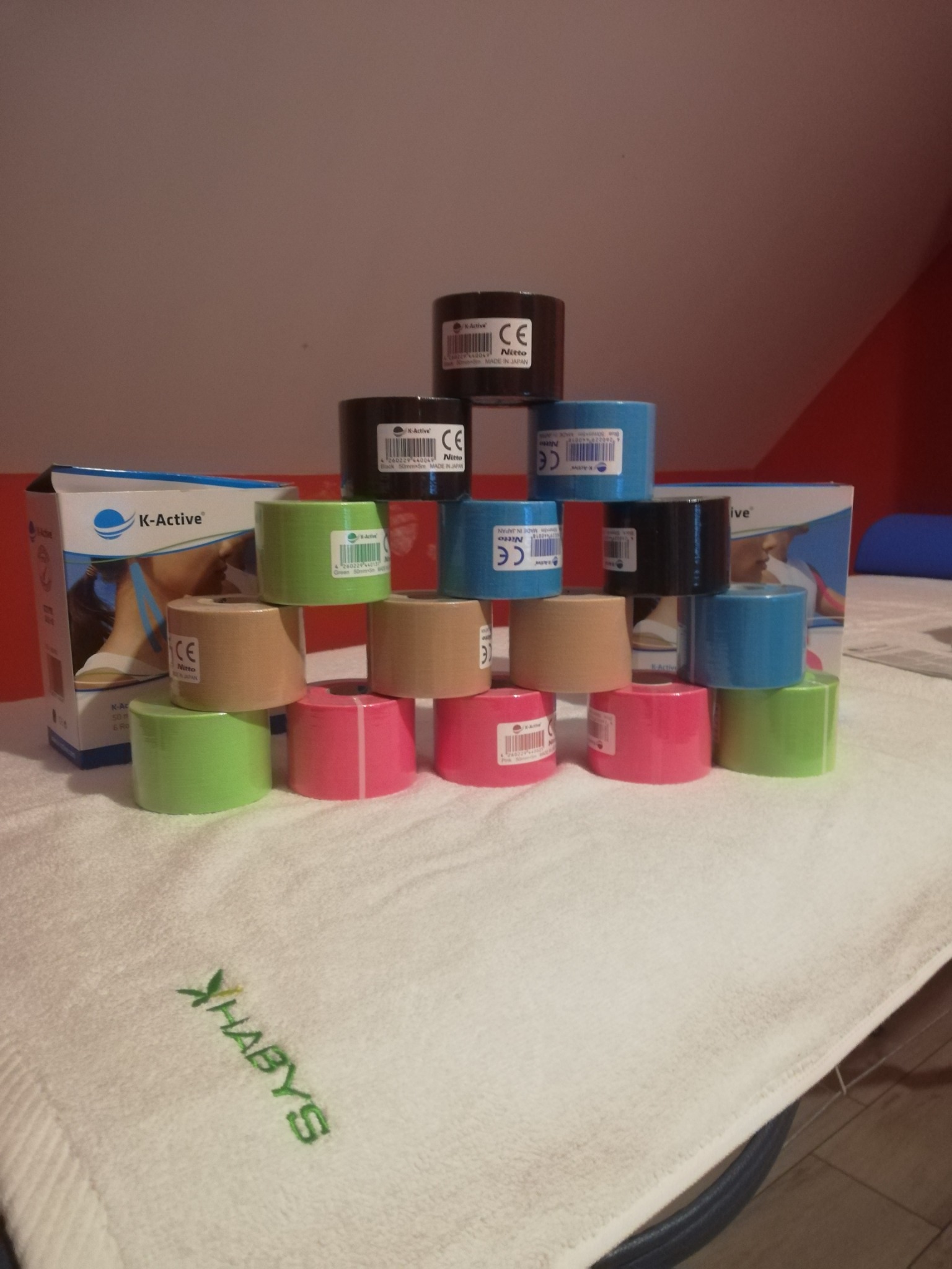 Piramida z kolorowych rolek taśmy K-Active Nitto na białym ręczniku z zielonym haftem logo XHABYS, z pudełkiem taśmy w tle.