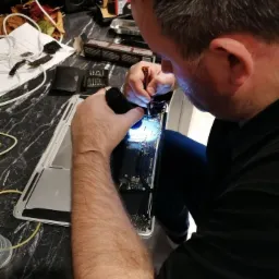 Technik przy biurku z czarnym blatem, podświetlając małą latarką płytę główną otwartego laptopa, precyzyjnie operuje śrubokrętem; w tle rozłożone narzędzia i akcesoria.