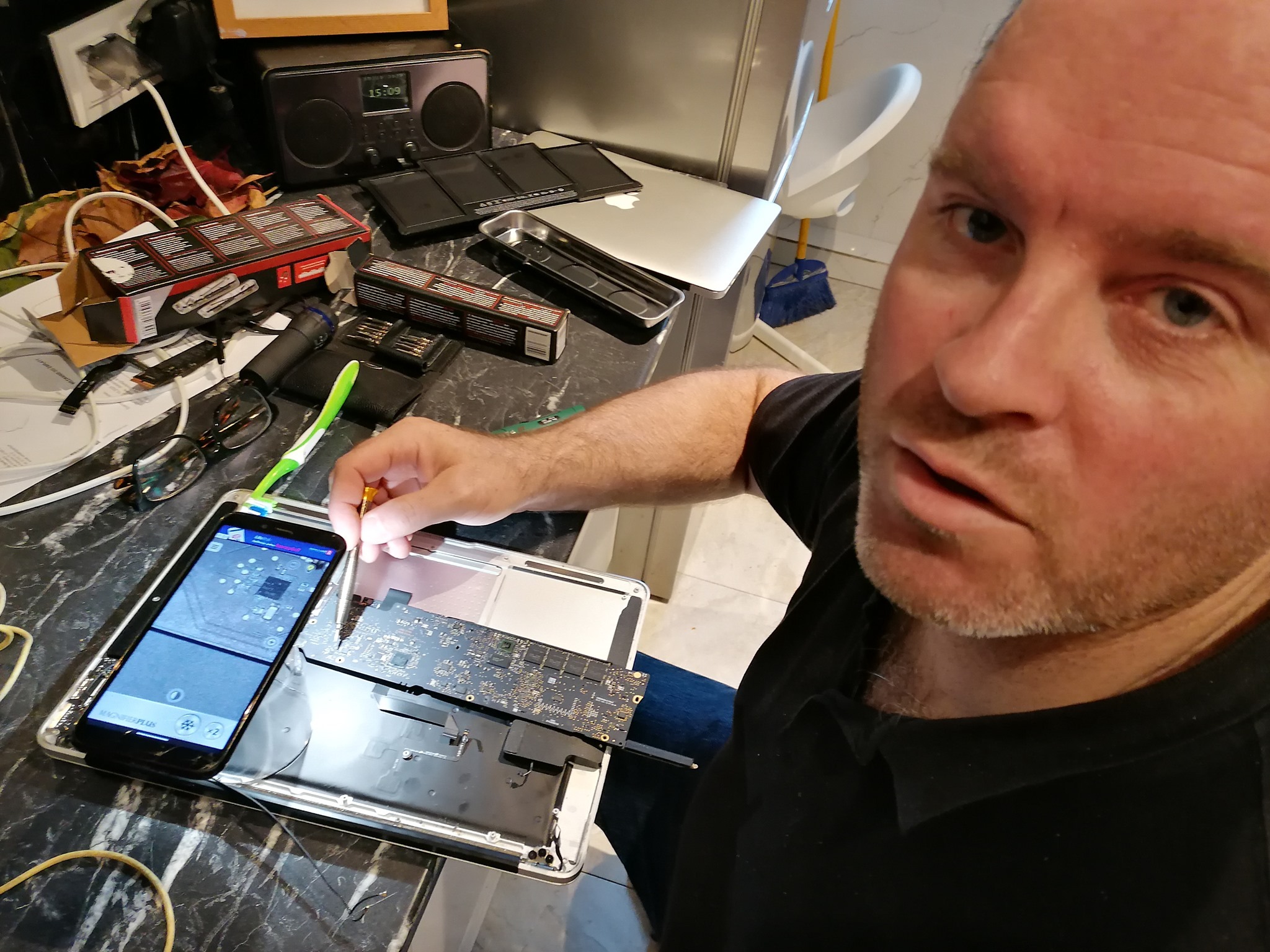 Mężczyzna z bliska, używający precyzyjnego śrubokręta do naprawy rozłożonego laptopa MacBook, z ekranem telefonu wyświetlającym powiększony widok płyty głównej i narzędziami w tle.