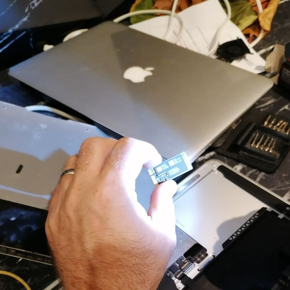 Mężczyzna trzyma w palcach moduł SSD nad rozłożonym MacBookiem w trakcie naprawy, widoczne śrubokręty i części komputera.