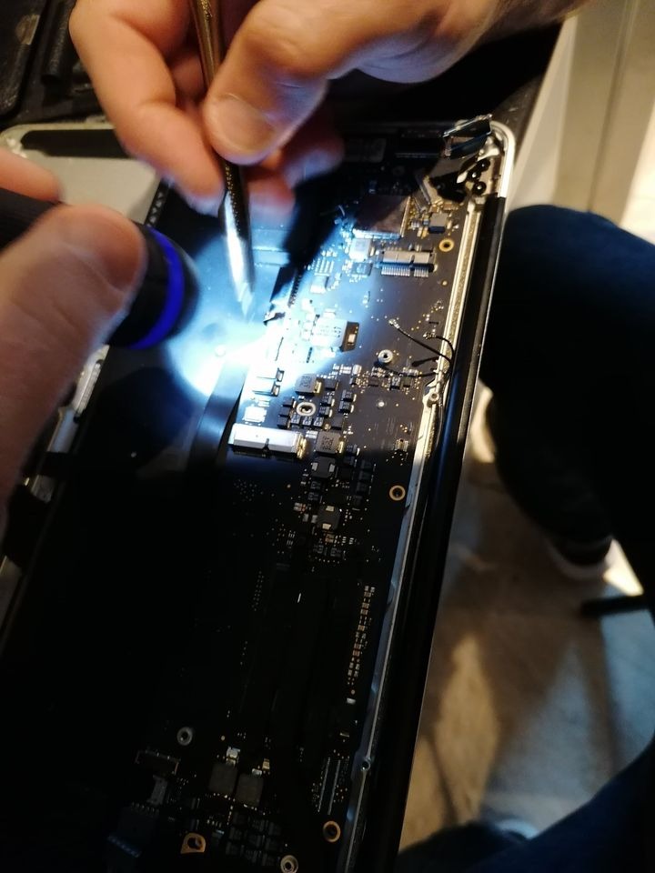 Otwarty laptop z widoczną płytą główną, oświetloną niebieską diodą LED, podczas diagnozy uszkodzenia za pomocą pęsety.