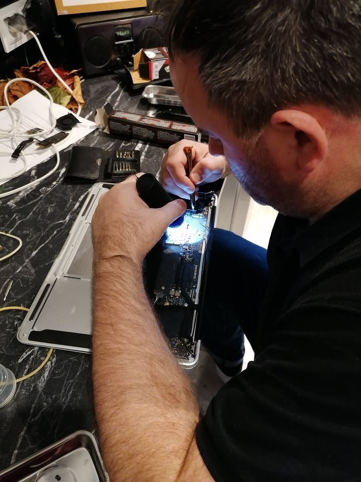 Technik przy biurku z czarnym blatem, podświetlając małą latarką płytę główną otwartego laptopa, precyzyjnie operuje śrubokrętem; w tle rozłożone narzędzia i akcesoria.