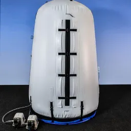 Biała komora hiperbaryczna z widocznymi czarnymi paskami i dwoma pompami powietrza u podstawy, na czarnym dywanie i tle błękitnej ściany.