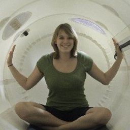 Hiperbaryczne Komory Tlenowe mHBOT Polska - Uśmiechnięta kobieta siedząca w pozycji lotosu wewnątrz białej komory hiperbarycznej, ręce uniesione w geście powitania, widoczne okna i oświetlenie wewnątrz komory.