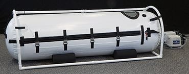 Biała komora hiperbaryczna z czarnymi pasami mocującymi, umieszczona na ramie z białych rur PCV, z widocznym urządzeniem sterującym po prawej stronie.